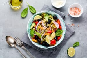 Whole grain pasta with vegetables on a white plate on a light grey slate, stone or concrete background. Top view with copy space